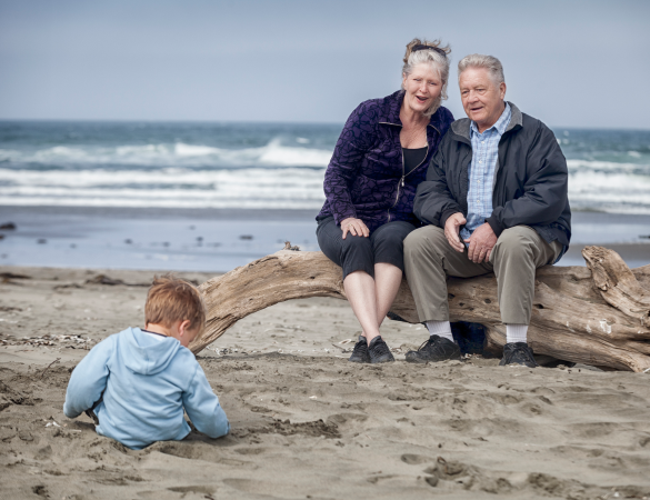 elderly couple sit on beach watching baby play in the sand