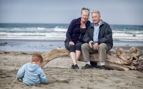 elderly couple sit on the beach watching a baby play in the sand
